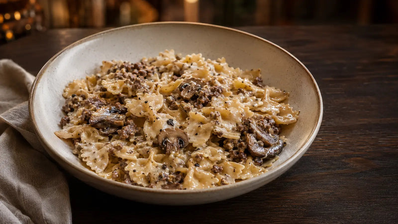 Una ciotola di pasta cremosa con carne macinata e funghi, servita su un tavolo di legno scuro con un tovagliolo piegato.
