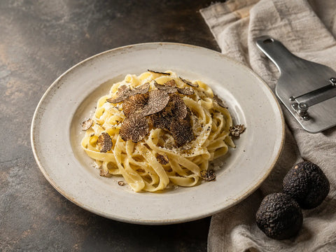Tagliatelle con scaglie di tartufo nero, tartufi interi e un affetta-tartufi su un piano di lavoro in pietra.